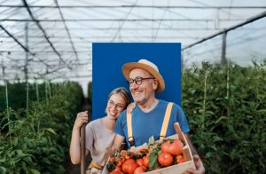 Ein Vater steht in einem Gewächshaus und hält einen Korb Gemüse. Seine Tochter lehnt ihren Kopf lachend an seine Schulter.