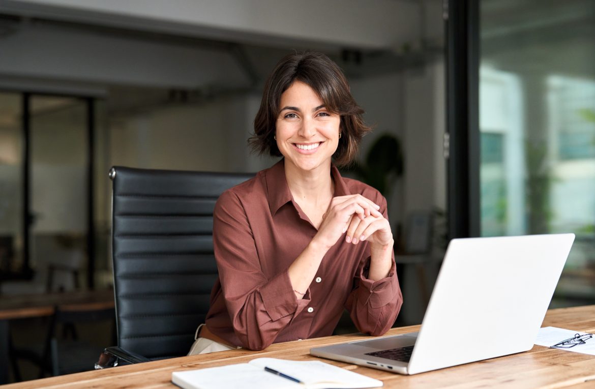 Lächelnde hispanische junge Unternehmerin, glückliche weibliche Führungskraft, die in die Kamera schaut und an ihrem Arbeitsplatz mit Laptop sitzt. Porträt einer selbstbewussten Geschäftsfrau in ihren 30ern am Schreibtisch.;