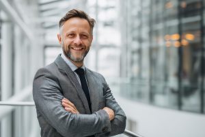 Ein Mann im gläsernen Gang eines Büros verschränkt lachend die Arme