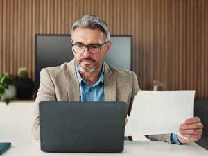 ein älterer herr sitzt mit einem blatt papierin der hand vor seinem laptop