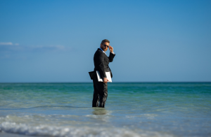 ein mann im anzug steht im meer mit laptop in der hand und setzt seine sonnenbrille auf.